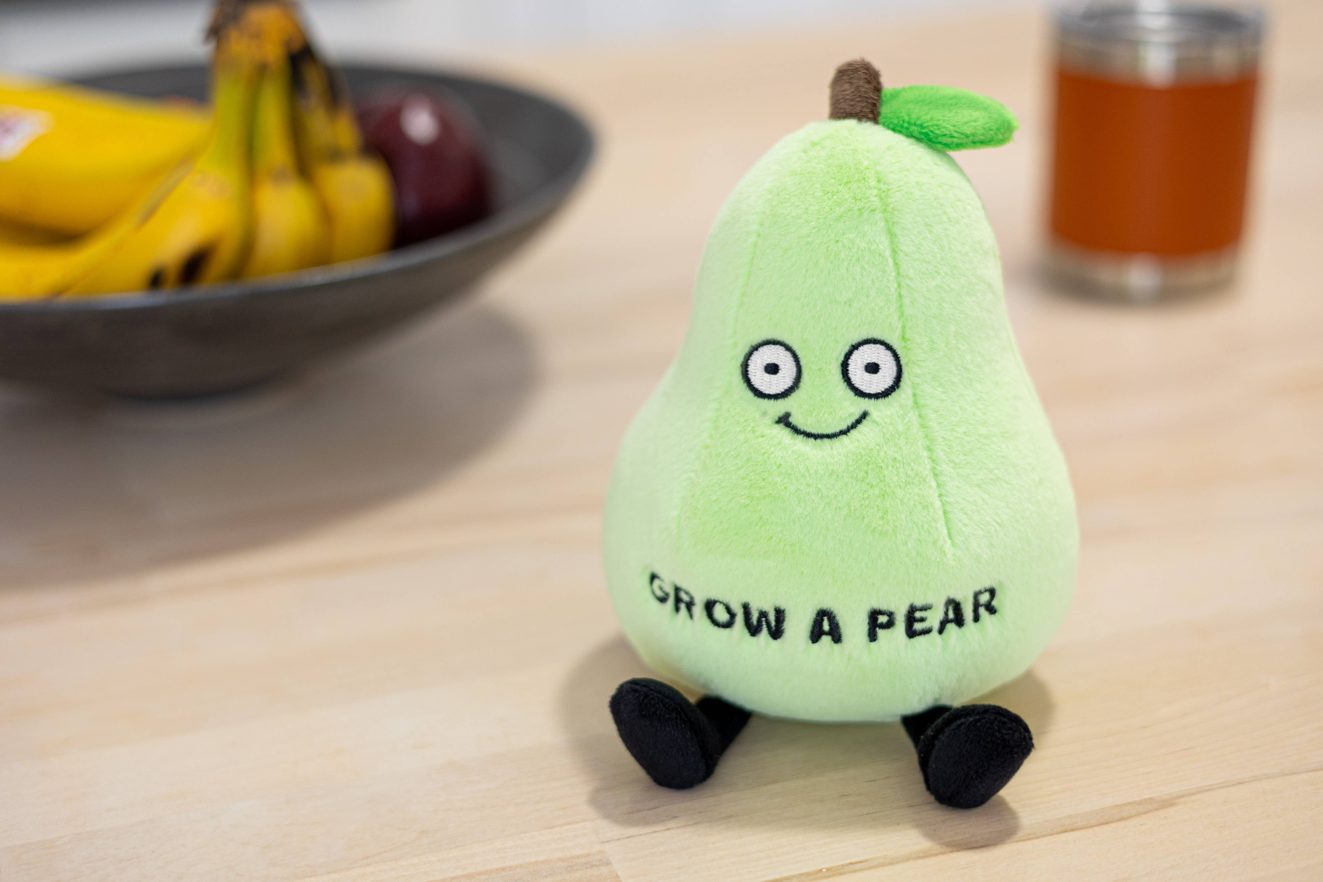 A green pear plushie with a smiley face, brown stem, and a green leaf on top. The front features black text that says "Grow a pear" and the plush has black legs sticking out.