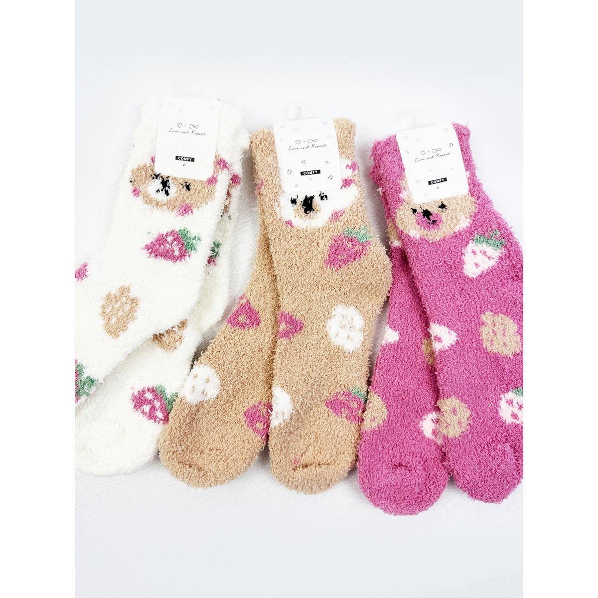 Three pairs of fuzzy, plush crew socks in white, light brown, and dark pink, featuring an embroidered bear face at the top cuff and a strawberry pattern.