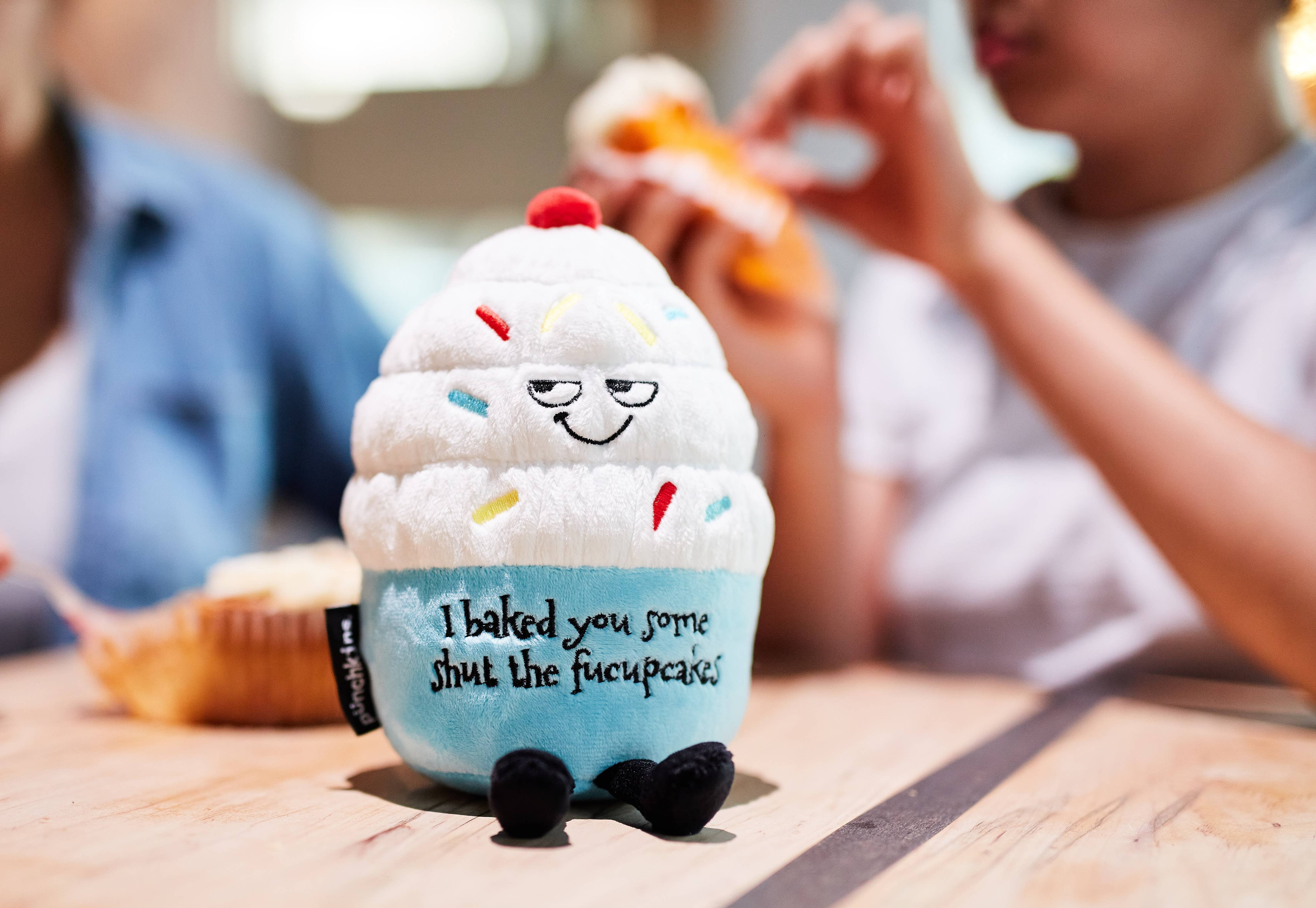 A cupcake plush with white frosting, rainbow sprinkles, and a blue base. The top features a sly, smiling face, while the bottom has black embroidered text that reads, "I baked you some shut the fucupcakes." Black legs sticking out the bottom and a red cherry on top.