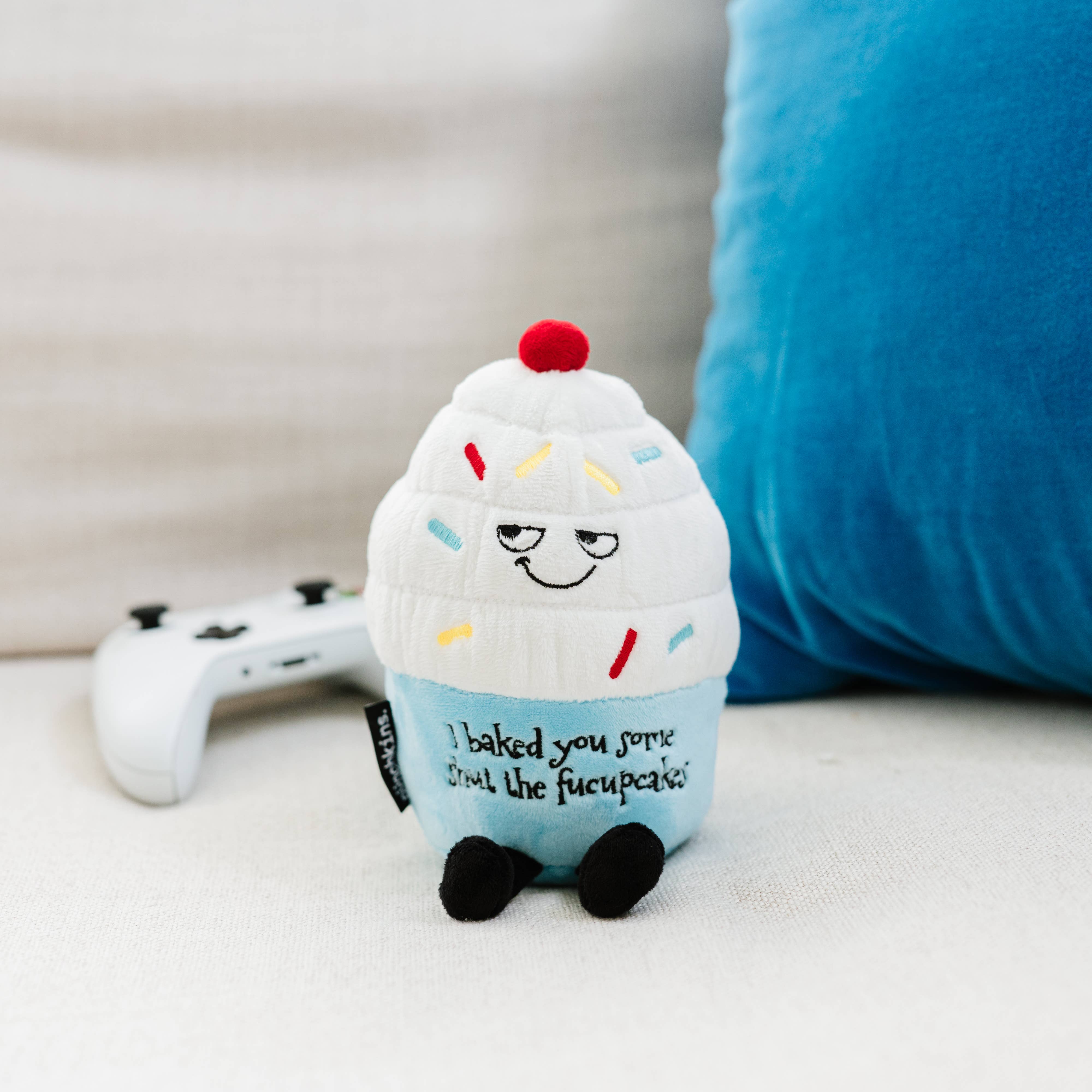 A cupcake plush with white frosting, rainbow sprinkles, and a blue base. The top features a sly, smiling face, while the bottom has black embroidered text that reads, "I baked you some shut the fucupcakes." Black legs sticking out the bottom and a red cherry on top.