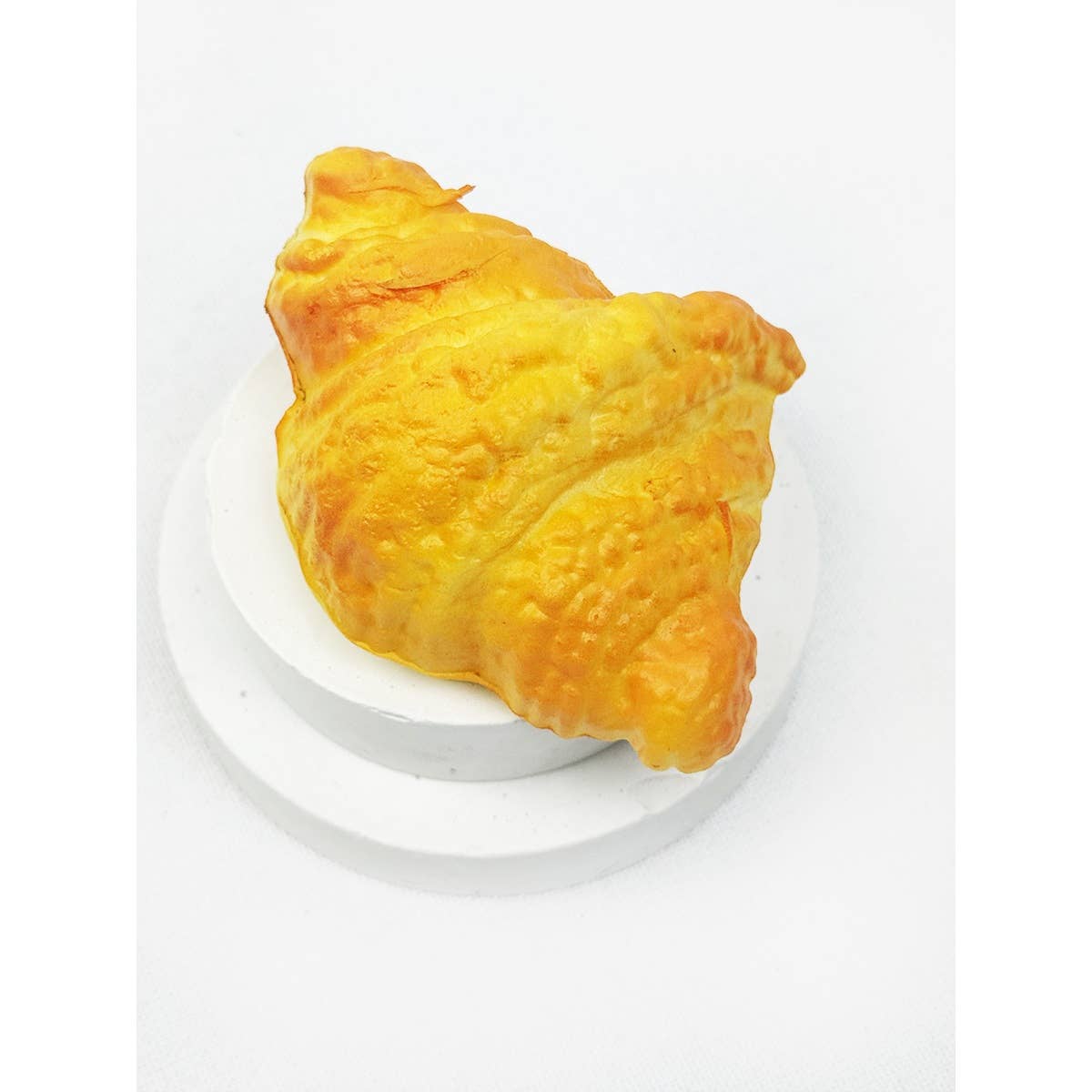 A realistic, golden-brown croissant-shaped slow rising bread squishy toy displayed on a small white circular platform.