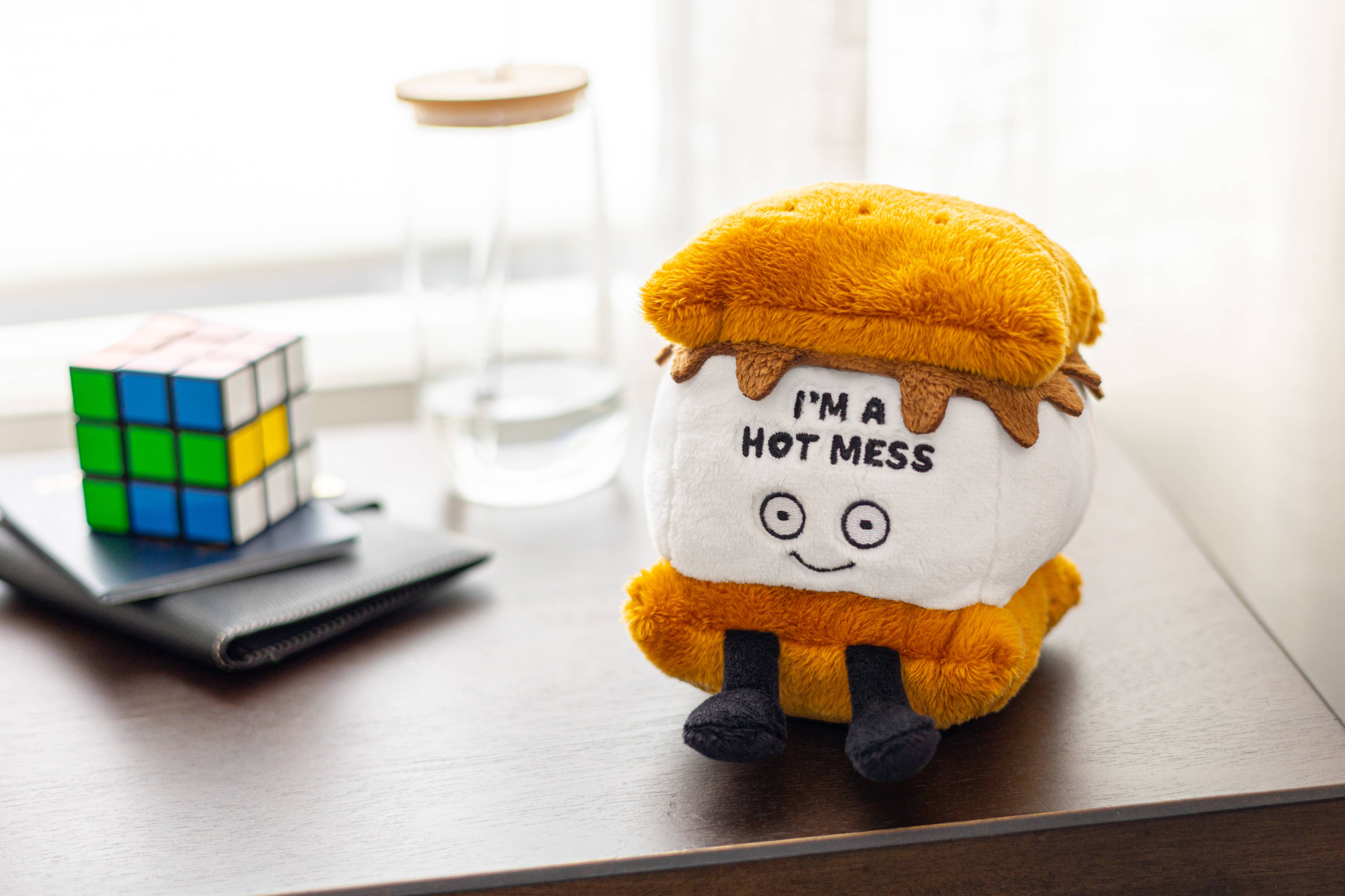 A s'more plushie featuring a smiley face, with black embroidered text that says 'I'm a hot mess.' The plushie has a layer of soft, golden-brown marshmallow, a piece of chocolate, and two graham crackers, all stitched together in a cute, playful design.