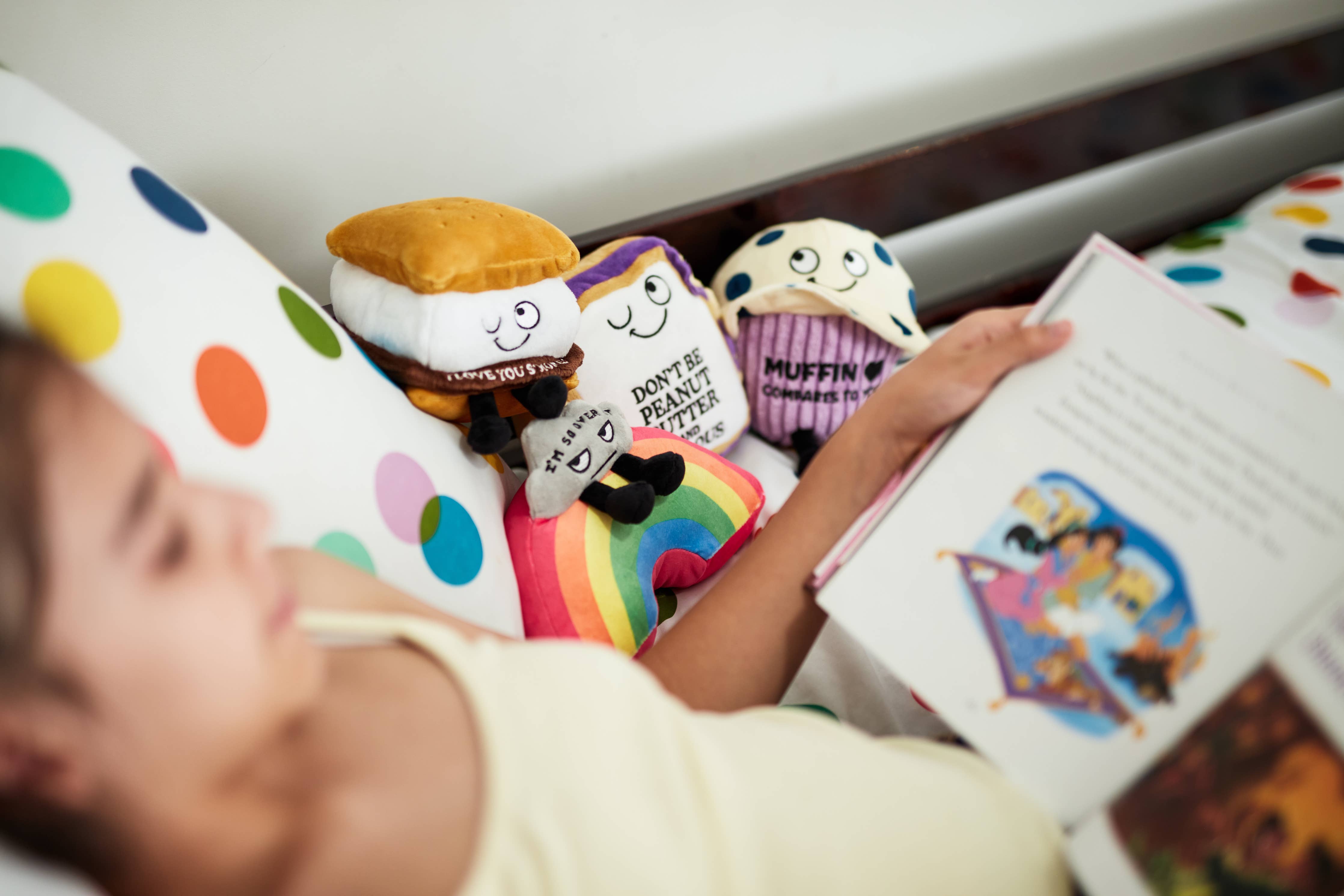 A plushie shaped like a s'more with a winky face. The chocolate layer features white lettering that reads "I love you s'more!" Pllushie is sitting on a bed with a child reading a book with other plushies.