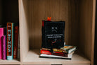 A black plush book sitting on a bookshelf with a sad expression and X-shaped eyes. White embroidered text reads, "I like my thrillers dark and twisted." A bloody knife detail is at the bottom, with black legs sticking out underneath and a red bookmark peeking from the top.