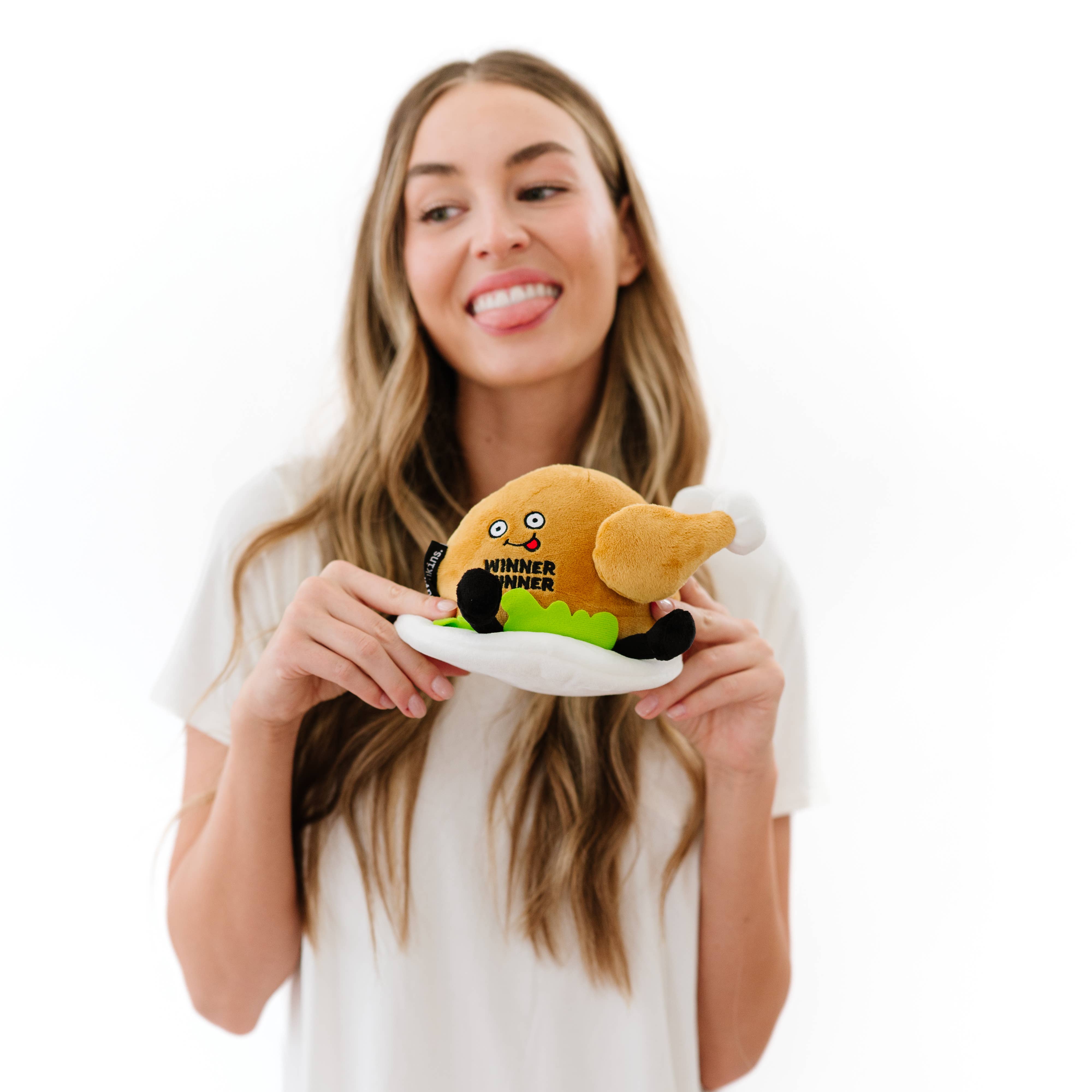 Woman sticking her tongue out while A cooked turkey dinner plush on a plate with green lettuce. The turkey has a smiley face with a red tongue sticking out and black text that says "Winner Winner." It also has black legs sticking out.