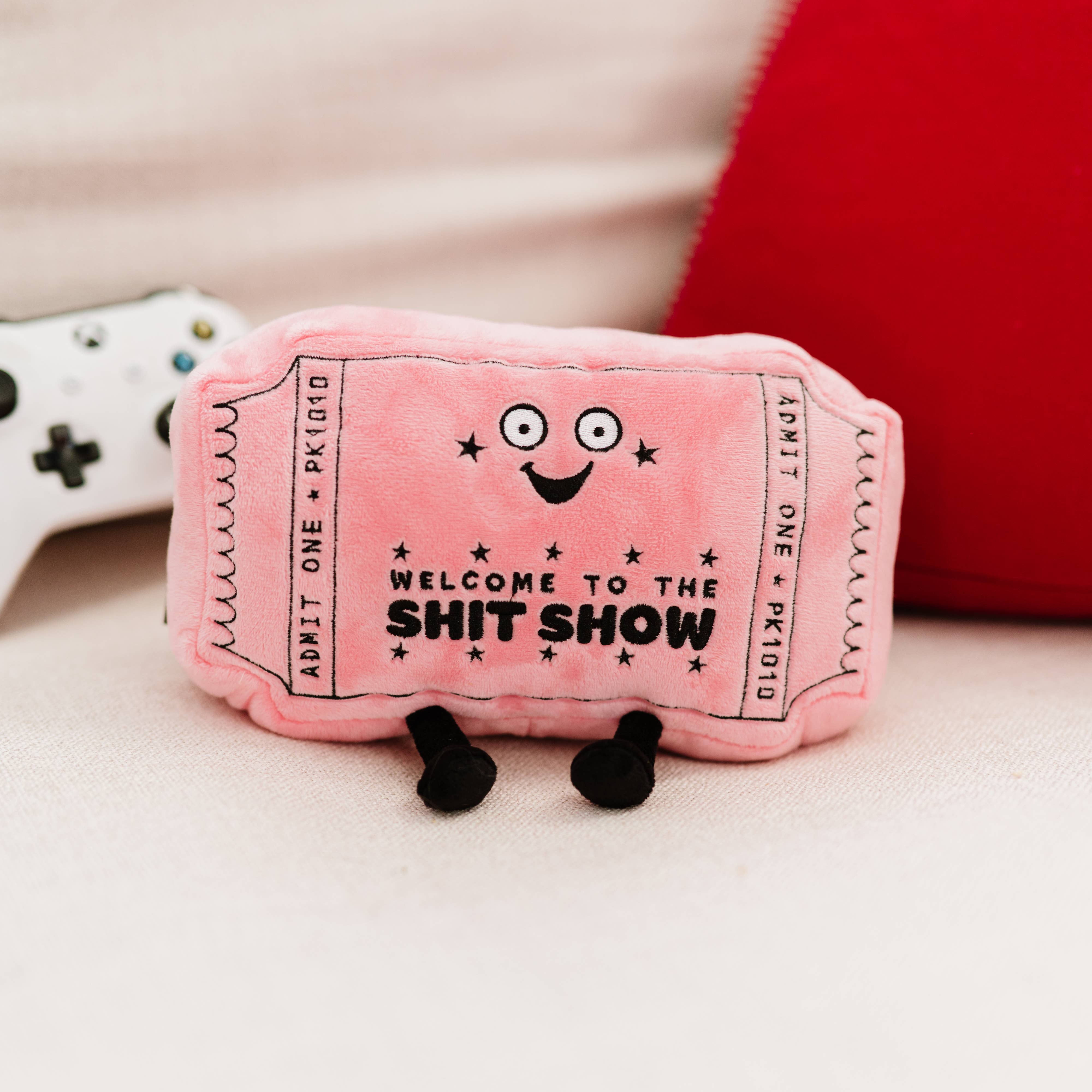 A pink plushie shaped like a movie ticket with a smiley face. It features black embroidery with the words "welcome to the shit show.