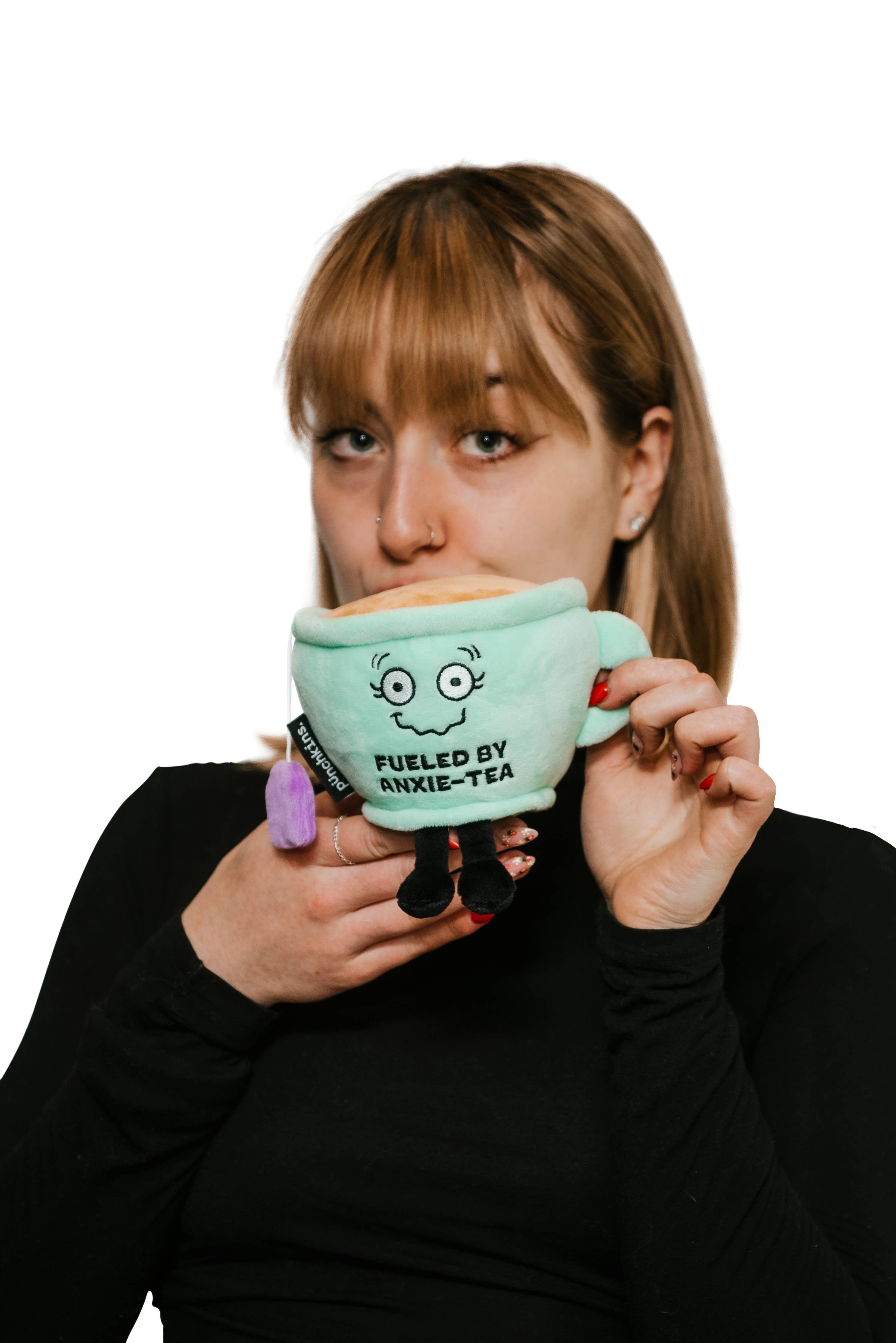 A green plush toy shaped like a smiling tea cup with a shaky smile. The cup is filled with soft, plush 'tea' on top, and a purple tea bag hangs from the side. The front displays the embroidered phrase "FUELED BY ANXIE-TEA" in bold letters. It has small black legs, a handle on one side, and an overall cute, cozy design.