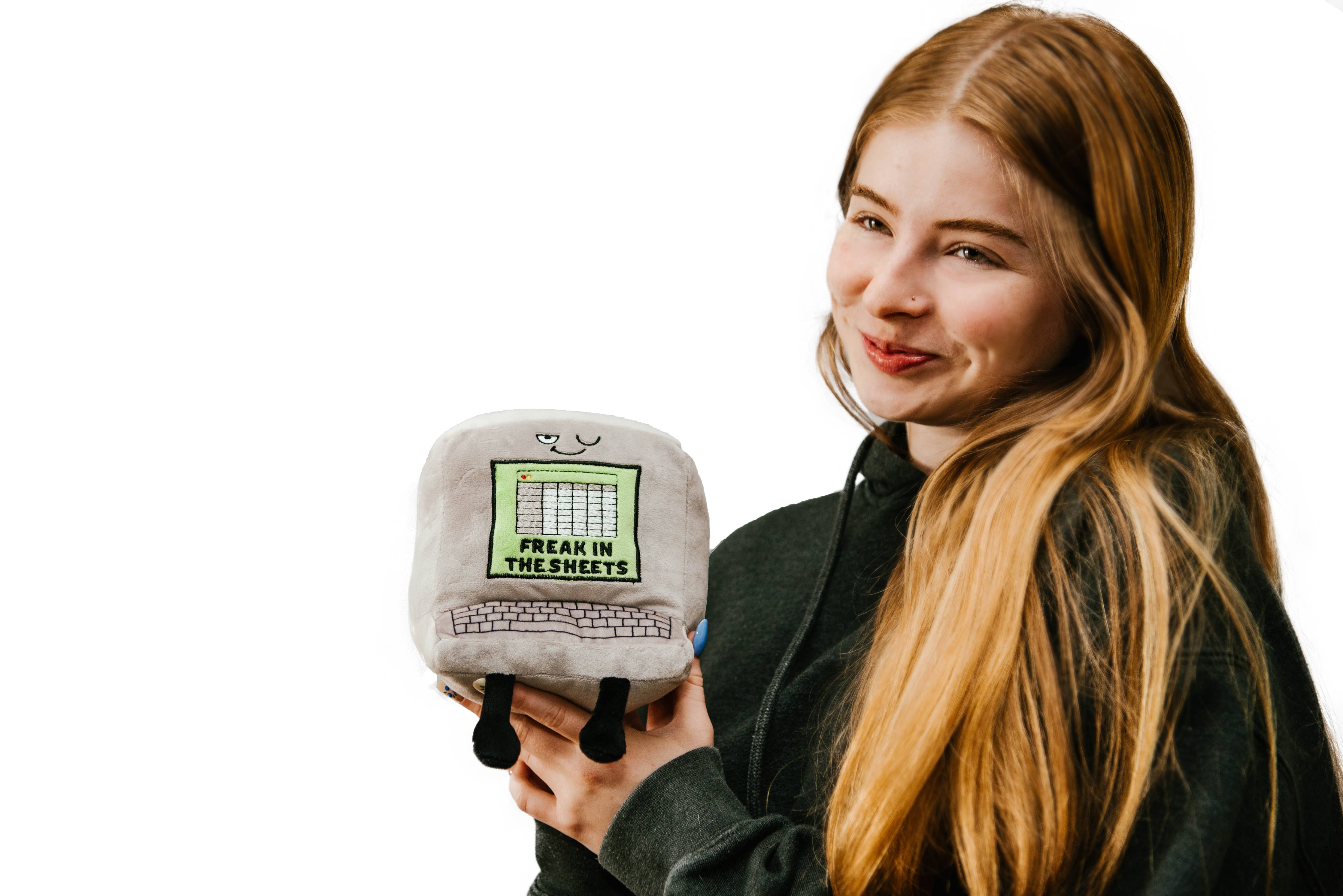 Woman holding a gray computer plush with a winky face, featuring a green Excel-style sheet embroidered with the words "Freak in the Sheets" in black. It also has an outlined keyboard design and black legs sticking out from the bottom.