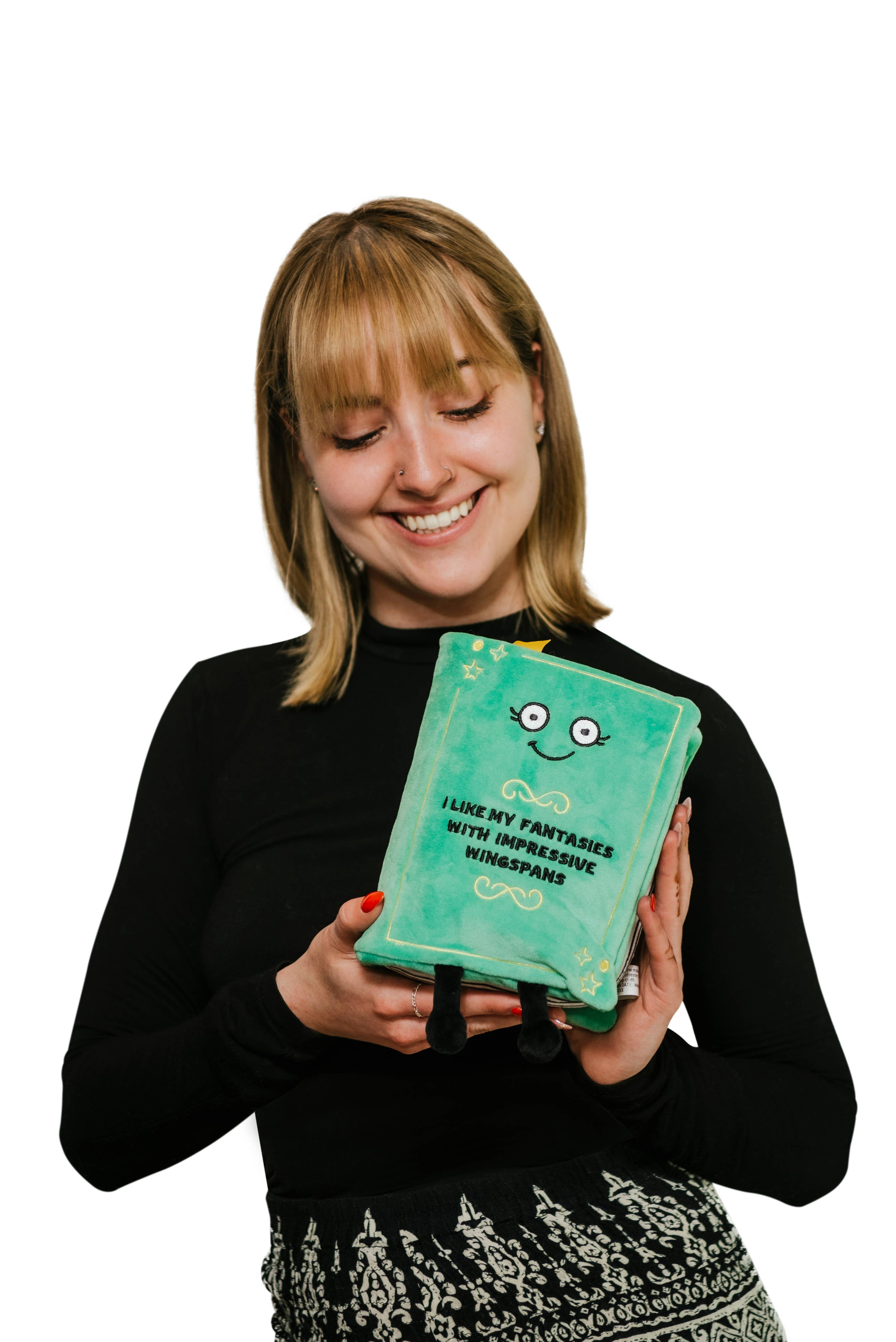 Woman holding A green plush with a smily face and embroidered black text that reads 'I like fantasies with impressive wingspans.' The plush has black legs sticking out and a yellow bookmark peeking from the top.