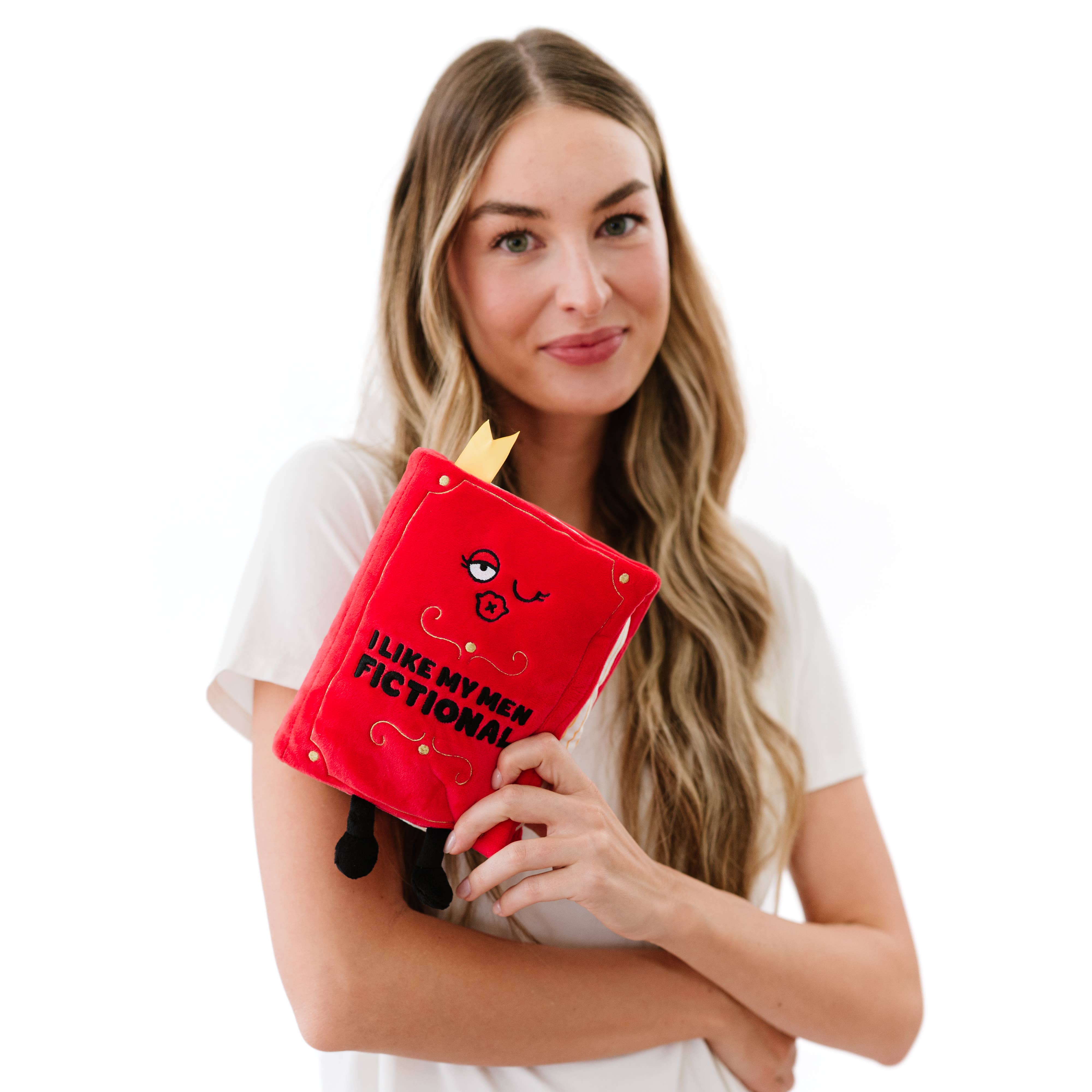 A red plush book with a kissy winky face and embroidered black text that reads 'I like my men Fictional.' The plush has black legs sticking out, a yellow bookmark peeking from the top, and 'A Spicy Romance' embroidered in black on the book's spine. A woman with long blonde hair, wearing a white shirt, is holding the plush.