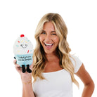 woman holding A cupcake plush with white frosting, rainbow sprinkles, and a blue base. The top features a sly, smiling face, while the bottom has black embroidered text that reads, "I baked you some shut the fucupcakes." Black legs sticking out the bottom and a red cherry on top.