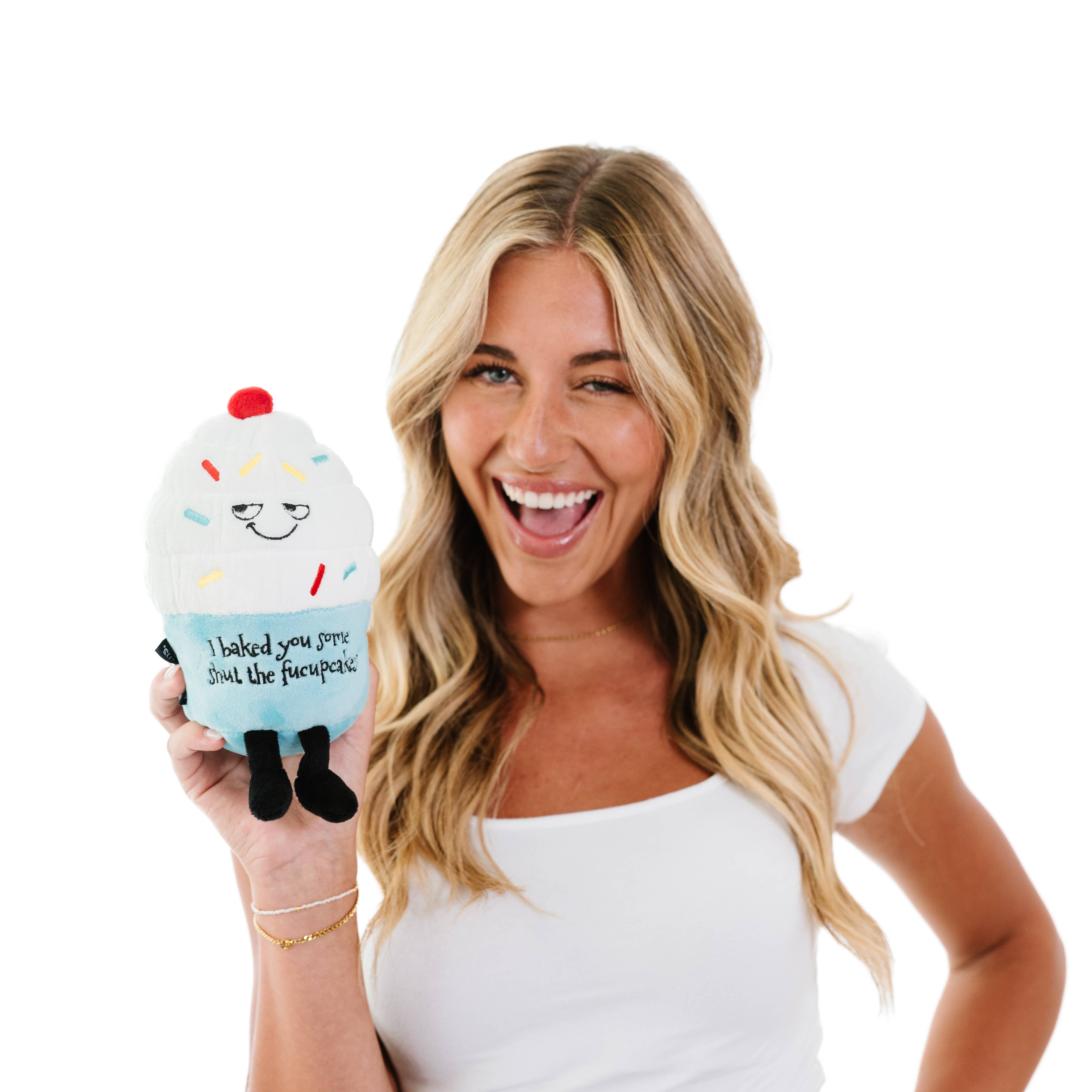 woman holding A cupcake plush with white frosting, rainbow sprinkles, and a blue base. The top features a sly, smiling face, while the bottom has black embroidered text that reads, "I baked you some shut the fucupcakes." Black legs sticking out the bottom and a red cherry on top.