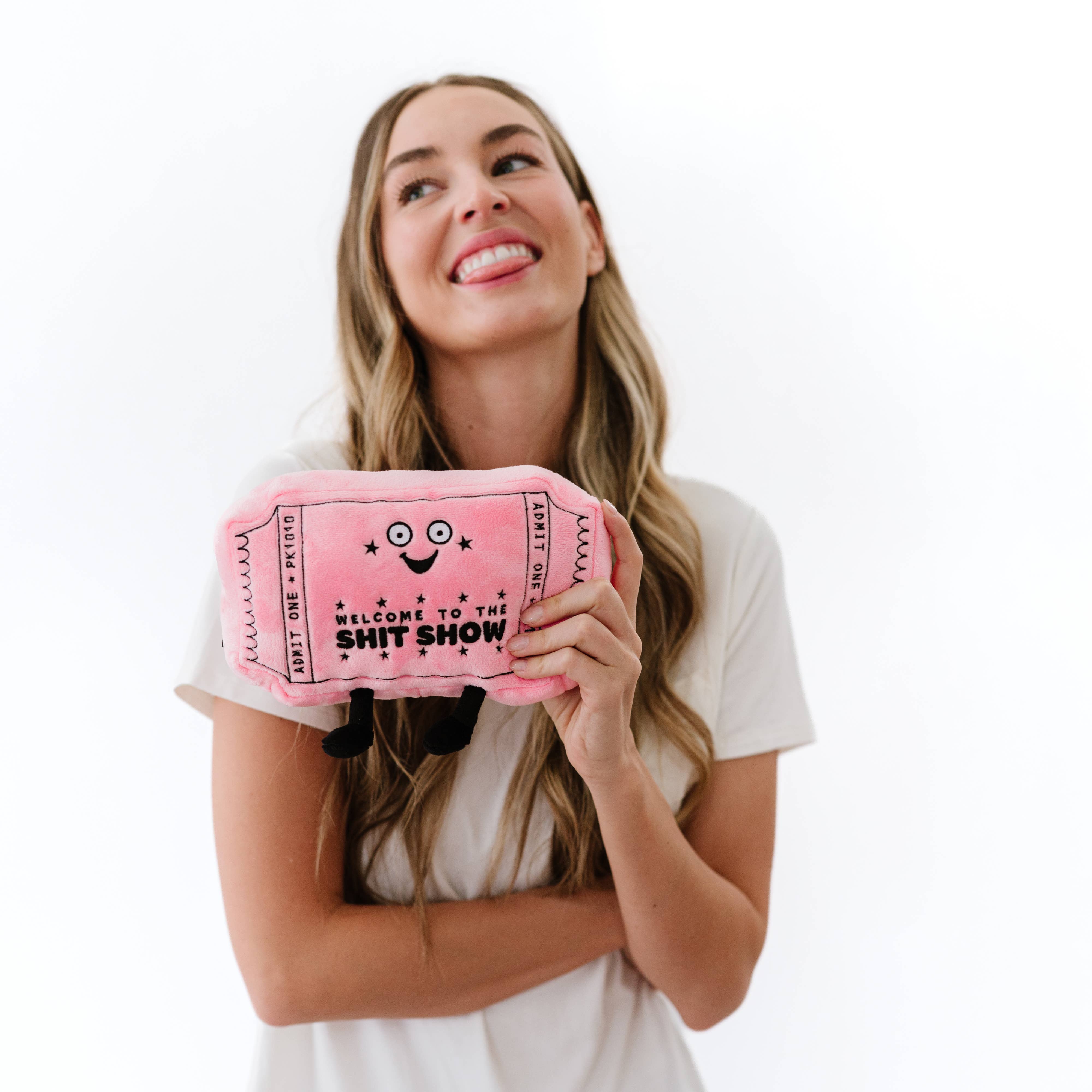 Woman holding a pink plushie shaped like a movie ticket with a smiley face. It features black embroidery with the words "welcome to the shit show.